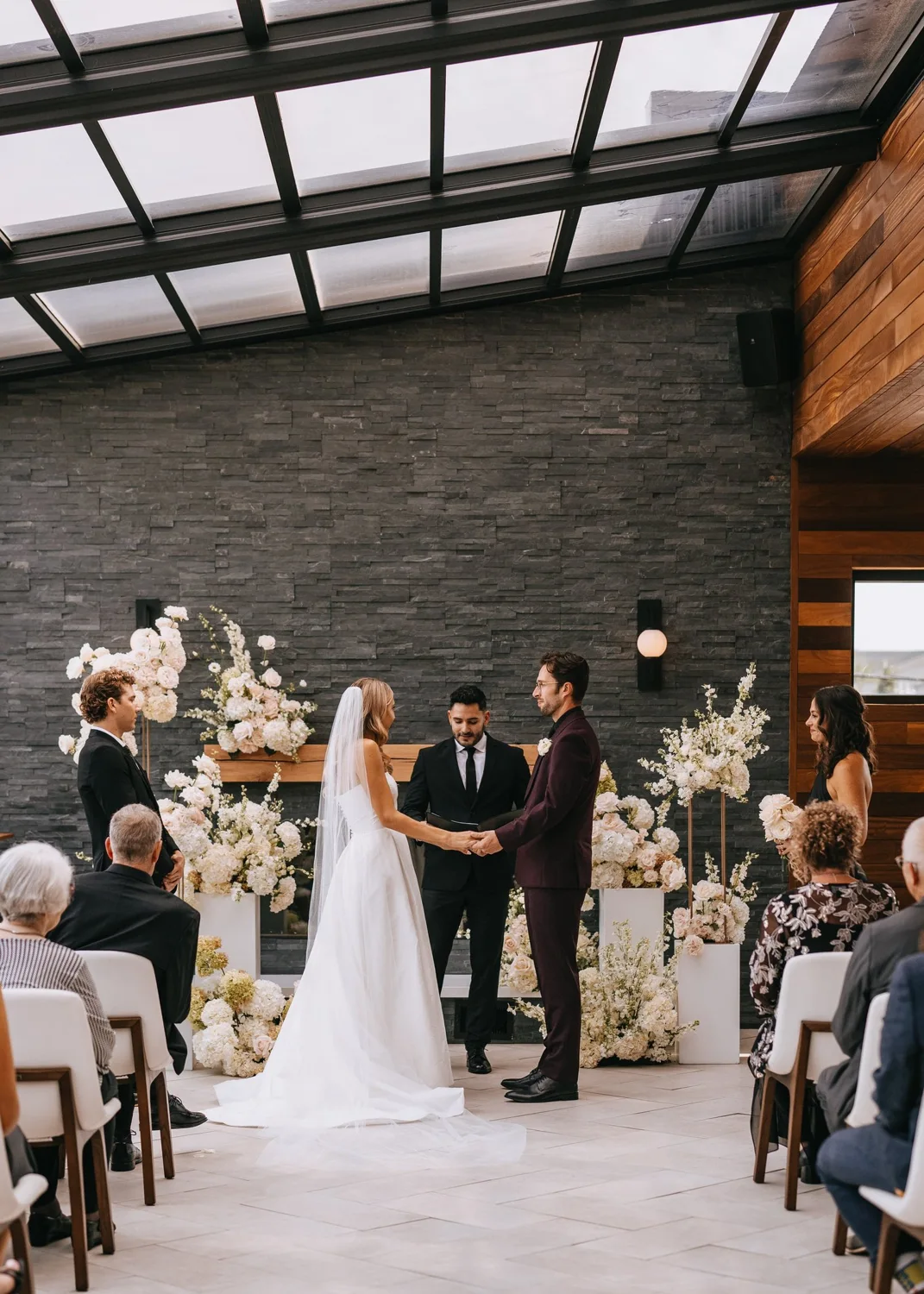 A couple exchanging vows beneath flowers in a glass-roofed wedding venue